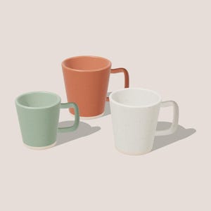May include: Three ceramic mugs with a speckled finish and a smooth, tapered design. One mug is sage green, one is terracotta orange, and one is off-white. Each mug features a matching handle and a solid, unglazed base rim.