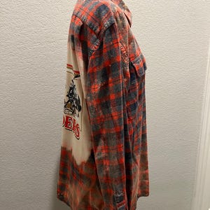 K&ouml;nnte beinhalten: Ein lang&auml;rmeliges, gekn&ouml;pftes Flanellhemd in einem rot-, grau- und schwarz karierten Muster. Auf der R&uuml;ckseite des Hemdes befindet sich eine Grafik mit dem Wort "Raiders" in Rot. Das Hemd wird auf einer schwarzen Schaufensterpuppe pr&auml;sentiert.