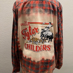 K&ouml;nnte beinhalten: Rot-graues Flanellhemd mit Karomuster und einem gebleichten Design auf dem R&uuml;cken. Das Design zeigt den Text "The Original Tyler Childers" mit einer Grafik einer Person, die ein Pferd reitet.