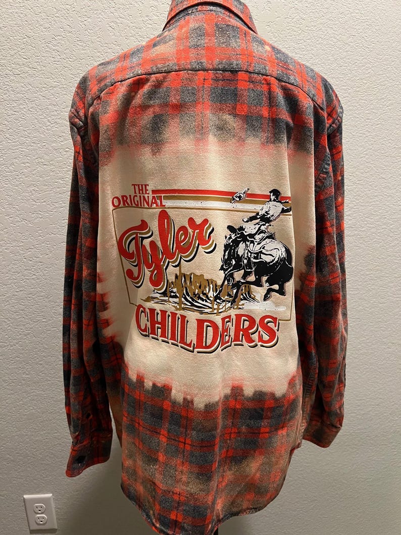 K&ouml;nnte beinhalten: Rot-graues Flanellhemd mit Karomuster und einem gebleichten Design auf dem R&uuml;cken. Das Design zeigt den Text "The Original Tyler Childers" mit einer Grafik einer Person, die ein Pferd reitet.