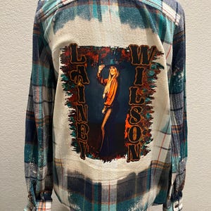 May include: A long-sleeved, button-down flannel shirt in a teal, navy, and white plaid pattern. The back features a bleached design with a graphic of a woman in a hat and the text "Lainey Wilson".