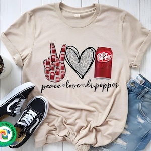 May include: A beige t-shirt with a peace sign, a heart, and a can of Dr Pepper. The text "peace love dr.pepper" is printed on the shirt.