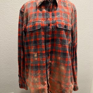 K&ouml;nnte beinhalten: Ein lang&auml;rmeliges Flanellhemd mit Kn&ouml;pfen in einem rot-, grau- und schwarz karierten Muster. Das Hemd hat zwei Brusttaschen und einen verwaschenen, gebleichten Effekt. Das Hemd wird auf einer schwarzen Schaufensterpuppe pr&auml;sentiert.
