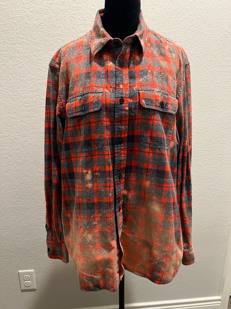 K&ouml;nnte beinhalten: Ein lang&auml;rmeliges Flanellhemd mit Kn&ouml;pfen in einem rot-, grau- und schwarz karierten Muster. Das Hemd hat zwei Brusttaschen und einen verwaschenen, gebleichten Effekt. Das Hemd wird auf einer schwarzen Schaufensterpuppe pr&auml;sentiert.