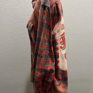 K&ouml;nnte beinhalten: Rot, schwarz und grau kariertes Flanellhemd mit einem gebleichten Design auf dem R&uuml;cken. Das Design enth&auml;lt den Text "THE GIN" in roter und schwarzer Schrift. Das Hemd hat zwei Brusttaschen und eine Knopfleiste.