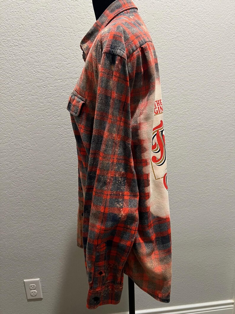 K&ouml;nnte beinhalten: Rot, schwarz und grau kariertes Flanellhemd mit einem gebleichten Design auf dem R&uuml;cken. Das Design enth&auml;lt den Text "THE GIN" in roter und schwarzer Schrift. Das Hemd hat zwei Brusttaschen und eine Knopfleiste.