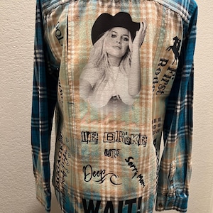 May include: A long-sleeved, button-down flannel shirt with a turquoise and black plaid pattern. The back features a black and white image of a woman wearing a cowboy hat, with text including "WE BROKE UP" and "WAIT!"