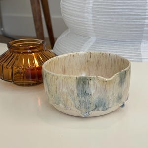 May include: A small, round ceramic bowl with a speckled cream base and a decorative glaze featuring vertical drips of muted blue and tan colors. The bowl has a slightly uneven rim with a small indentation on one side, creating a subtle spout.