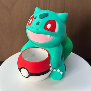 Può includere: Una statuetta di un personaggio Pokemon stampata in 3D di colore verde acqua e rosso che tiene una Pokéball rossa e bianca.
