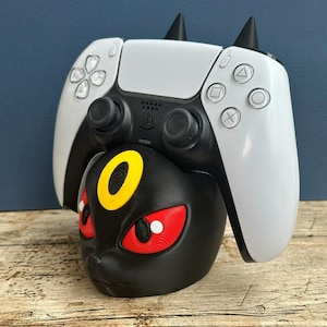 Op de afbeelding: Een zwarte Umbreon-thema controllerstandaard met een witte en zwarte PlayStation 5 controller. De standaard heeft rode ogen en een gele ring. De controller heeft zwarte joysticks en knoppen.