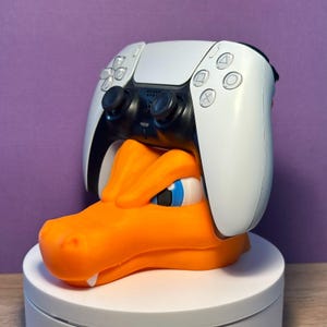 Może przedstawiać: Biało-czarny kontroler PlayStation 5 jest zamontowany na pomarańczowej, trójwymiarowej podstawce w kształcie głowy smoka. Podstawka stoi na białym, okrągłym cokole.