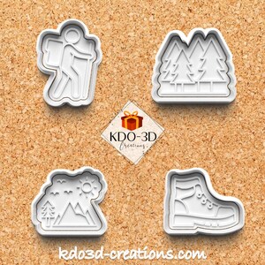 Op de afbeelding: Vier witte plastic koekjessnijders met buitenthema's. Ontwerpen omvatten een wandelaar met een rugzak, twee dennenbomen, een berglandschap met bomen en zon, en een wandelschoen. De website kdo3d-creations.com is ook zichtbaar.