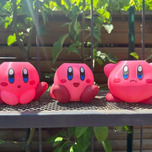 Può includere: Tre figurine di personaggi rosa con diverse espressioni facciali. Le figurine hanno la forma di un personaggio rosa con occhi grandi e una bocca piccola. Le figurine sono sedute su uno scaffale di metallo nero.