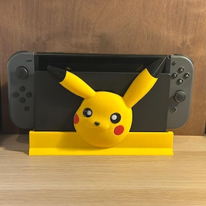 Könnte beinhalten: Ein gelber Pikachu-Stand für eine Nintendo Switch-Konsole. Der Ständer ist aus 3D-gedrucktem Kunststoff gefertigt und verfügt über einen gelben Pikachu-Kopf mit schwarzen Ohren und roten Wangen. Der Ständer hat eine gelbe Basis und ist so konzipiert, dass er die Switch-Konsole aufrecht hält.