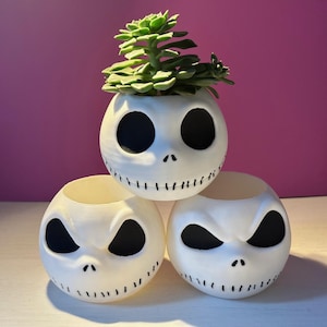 Könnte beinhalten: Drei weiße Keramik-Blumentöpfe in Form von Jack Skellingtons Gesicht aus dem Film Nightmare Before Christmas. Der obere Topf hat eine kleine grüne Sukkulente, die darin wächst.