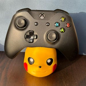 Può includere: Un controller Xbox nero con pulsanti gialli, blu e verdi è seduto su una statuetta di Pikachu gialla.