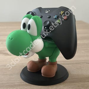 Può includere: Un supporto per controller a tema Yoshi verde che tiene un controller Xbox nero. Il supporto presenta una figura di Yoshi verde con dettagli bianchi, scarpe marroni e una base nera. Il testo "3dartcadeux.etsy.com" è visibile.