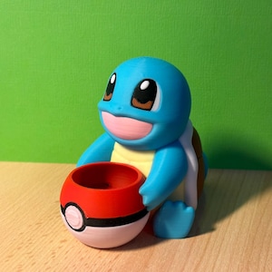 Peut inclure: Une figurine de Squirtle Pokémon bleue et blanche tenant un Poké Ball rouge et blanc. Le Poké Ball a une bande noire au milieu.