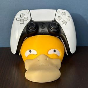 Op de afbeelding: Een witte PlayStation 5 DualSense-controller zit in een gele en beige 3D-geprinte standaard in de vorm van een eend.