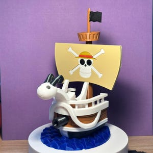 Może przedstawiać: Szczegółowy model statku z żaglem z czaszką i skrzyżowanymi piszczelami. Statek ma biało-brązowy kadłub, czarną flagę i białą głowę barana. Żagiel przedstawia czaszkę w słomkowym kapeluszu. Statek stoi na niebieskiej podstawie.