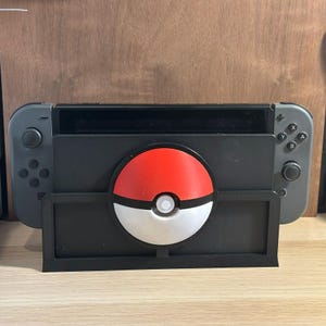 Peut inclure: Un support noir pour console Nintendo Switch avec un design de Poké Ball rouge et blanc. Le support est en plastique et possède une fente pour accueillir la console. La Poké Ball est un objet populaire de la franchise de jeux vidéo Pokémon.