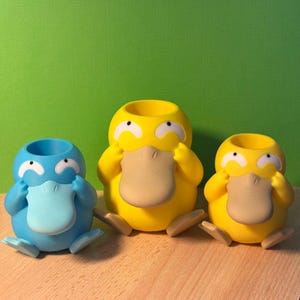 Op de afbeelding: Drie kleine plastic figuurtjes van een cartoon-eendensnaveldier. De eendensnaveldieren staan in een rij, met de blauwe links, de gele in het midden en de gele rechts. Alle drie eendensnaveldieren hebben hun handen voor hun ogen en kijken weg van de camera.