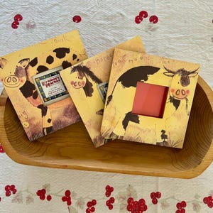 May include: Three farm animal-themed picture frames in a wooden tray. The frames feature illustrations of a cow, horse, and pig on a yellow background. One frame has a small sign that reads "Framer's Market". The frames are on a white tablecloth with red berry designs.