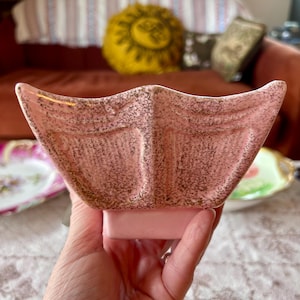 May include: A pink ceramic planter shaped like a whale tail with a speckled gold finish. The object features a wide, curved top edge and a solid rectangular base. The surface has a textured appearance with a light pink base color and gold flecks.