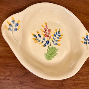 May include: Cream-colored ceramic serving dish with a floral design. The dish has a slightly irregular shape with two handles. The floral design features blue, yellow, and red flowers with green leaves. The dish is on a wooden surface.