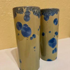 May include: Two ceramic tumblers with a light tan base color and blue crystalline patterns. The rims are a darker blue with a textured appearance. The tumblers are cylindrical and appear to be handmade.