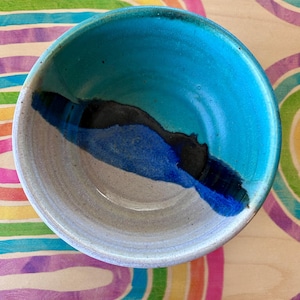 May include: A handmade ceramic bowl with a turquoise interior and a gray exterior. The bowl features a dark blue and black accent design. The bowl is sitting on a rainbow-colored surface.