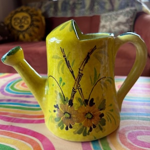 May include: A yellow ceramic watering can with a green handle and spout. The watering can is decorated with hand-painted floral and bamboo designs in shades of brown, green, and yellow. The watering can is sitting on a colorful, rainbow-patterned surface.