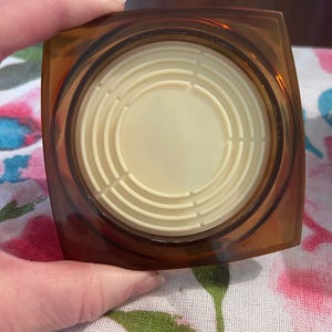 May include: A vintage, square-shaped compact with a translucent amber-colored exterior. The interior features a cream-colored, circular design with concentric rings. The compact is held by a hand, suggesting its portable nature and cosmetic use.