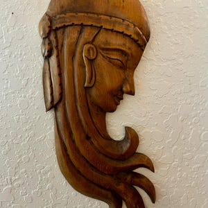 May include: Wooden wall art depicting a woman's profile. The sculpture is carved from wood and features flowing hair and a serene expression. The piece is a warm brown color and is mounted on a textured white wall.