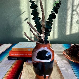 May include: A ceramic vase with a black and white design, holding eucalyptus branches and dried stems. The vase sits on a rectangular, gold-toned tray. Books and a woven basket are in the background, on a colorful striped textile.