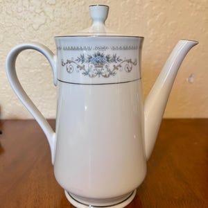May include: A white porcelain coffee pot featuring a delicate floral pattern with blue roses and brown scrollwork accents. The pot includes a matching lid with a small knob, a curved handle, a tapered spout, and thin silver trim along the edges.