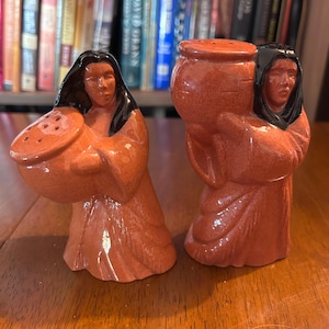 May include: A pair of ceramic salt and pepper shakers. Each shaker is a terracotta-colored figure of a person with black hair and a draped garment. One holds a bowl with holes, the other a pot. The figures are standing.