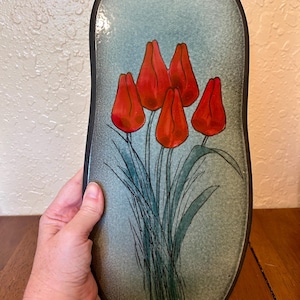 May include: An elongated oval-shaped decorative tray featuring a speckled light blue and grey background with a central illustration of five vibrant red tulips with green stems and leaves, all enclosed within a dark brown or black rimmed border.