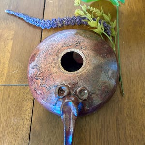 May include: A round, flat ceramic oil lamp with a metallic, iridescent bronze and purple glaze. The surface features etched wave patterns and circular motifs, with a central circular opening and a protruding spout at the front edge.