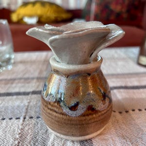 May include: A small ceramic bud vase featuring a rounded base with a glossy brown, tan, and blue drip glaze finish. The top of the vase is shaped like an open flower with light gray, smooth petals that extend outward from the center opening.