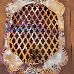 May include: An oval-shaped metal trivet with an iridescent, tarnished finish and a central diamond lattice cutout pattern. The frame features ornate floral relief detailing at the top and bottom edges, with a small circular maker's mark sticker.