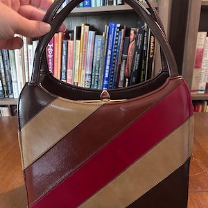May include: A structured handbag featuring a diagonal striped pattern in shades of dark brown, tan, and deep red. The bag has a glossy finish, a single arched handle, and a gold-toned metal clasp closure at the top center of the main compartment.