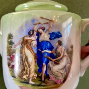 May include: A white porcelain teapot with a light green lid and handle. The teapot features a painted scene of three women in flowing gowns, with a bow and arrows. The women are in shades of blue, brown, and pink.
