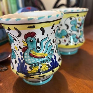 May include: Two hand-painted ceramic vases with a rooster design. The vases are white with blue, yellow, and red accents. The rooster is blue with a red comb. The vases have a flared shape and are approximately 4 inches tall.