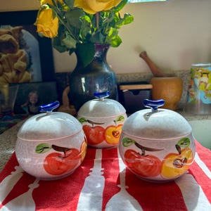 May include: Three ceramic jars with lids, each adorned with a hand-painted apple design in red and yellow, sit on a red and white striped cloth. The jars have blue handles and are arranged in front of a vase of yellow roses.