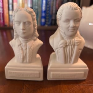 May include: Two off-white ceramic busts of historical figures. The busts are on rectangular bases with engraved text. One bust depicts a man with long hair and the other a man in a suit with a bow tie. The background includes a bookshelf.