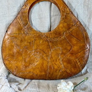 May include: A handcrafted, brown leather handbag with a large circular handle. The bag is constructed from irregularly shaped leather pieces, stitched together with dark thread. The bag's overall shape is oval, with a stitched border.