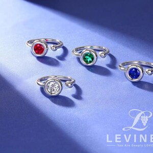 May include: Four silver rings with different colored gemstones. The rings have a simple design with a round stone set in a bezel setting. The stones are red, green, blue, and clear.