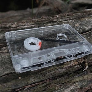 Könnte beinhalten: Ein transparentes Plastik-Kassettenband mit einem roten und weißen Etikett. Das Band liegt auf einem Stück verwittertem Holz.