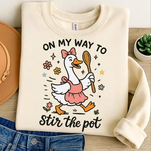 May include: Cream-colored sweatshirt with a whimsical graphic of a goose wearing a pink apron and holding a wooden spoon. Text reads "ON MY WAY TO STIR THE POT" in black script, surrounded by flowers and stars.